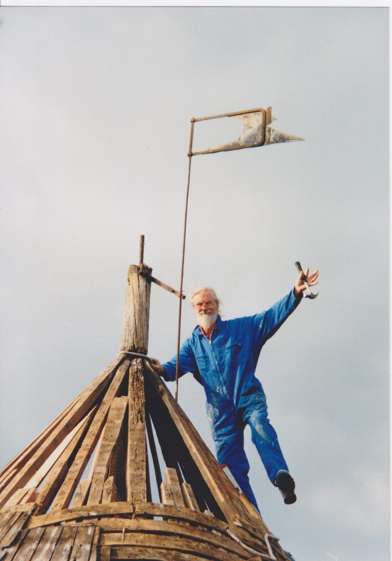 Brian Chantry, en bleu de travail, debout sur la charpente nue de la toiture de la tour, marteau à la main